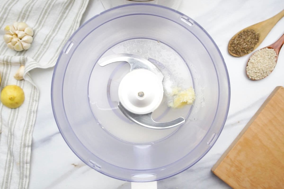 Close-up of food processor with garlic, lemon, and butter on marble countertop.