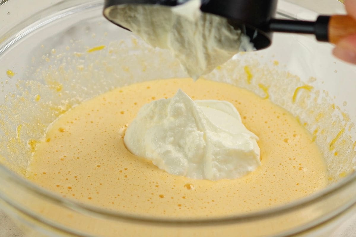 A close-up of a hand pouring batter into a mixing bowl with whipped cream for baking.
