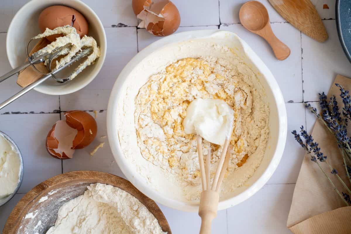 Close-up of flour, eggs, and butter for baking, with cracked eggshells and utensils on a rustic kitchen table.