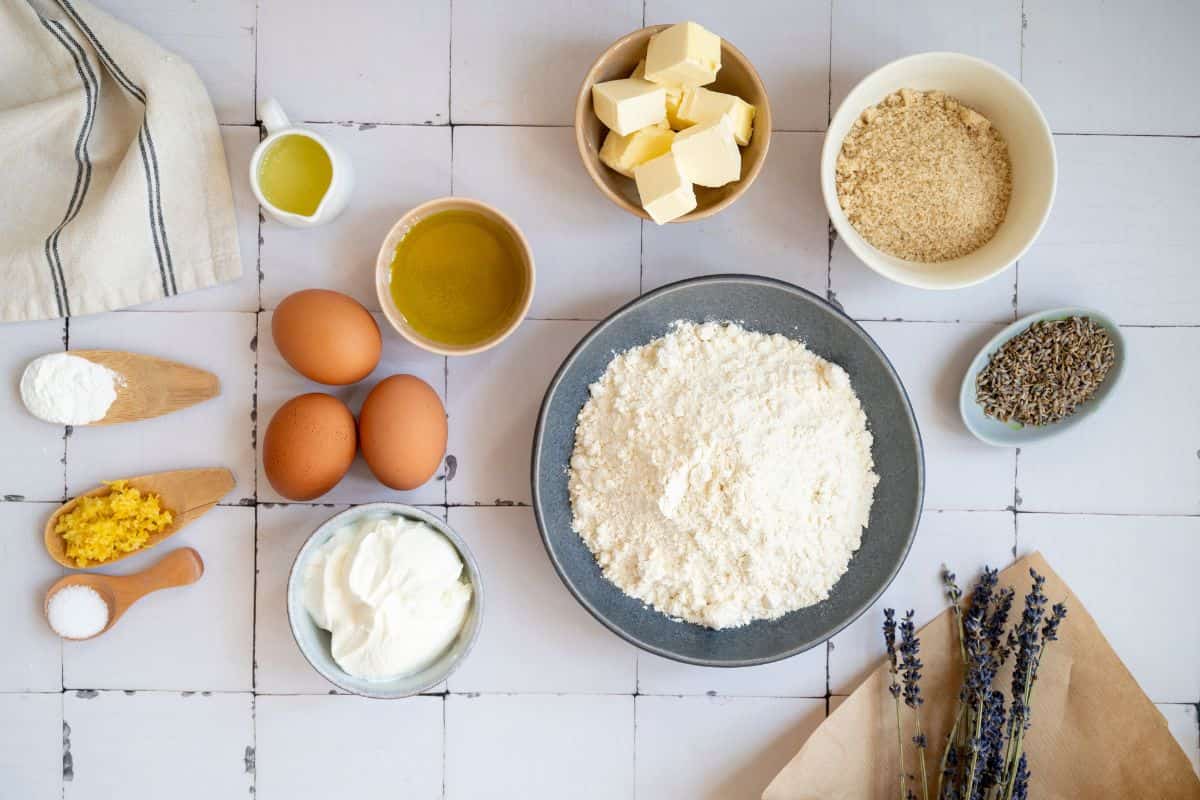 Fresh ingredients including flour, eggs, butter, and lavender on a rustic white table.