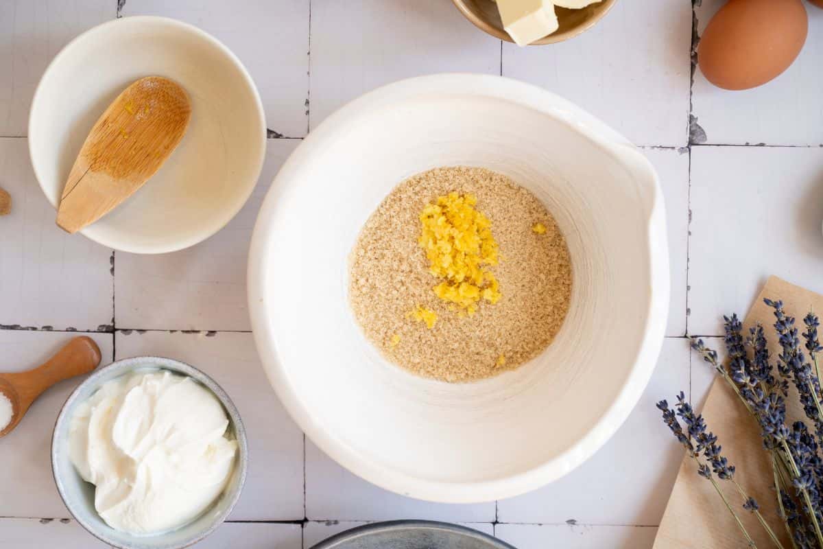 A white mixing bowl with butter and sugar, surrounded by eggs, flour, and lavender on a white tiled surface.