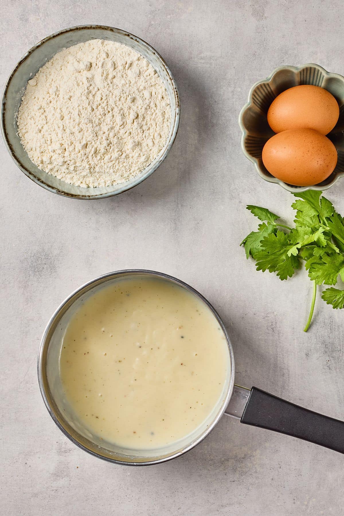 Bowl of flour, eggs in a small dish, milk in a pan, and fresh cilantro.