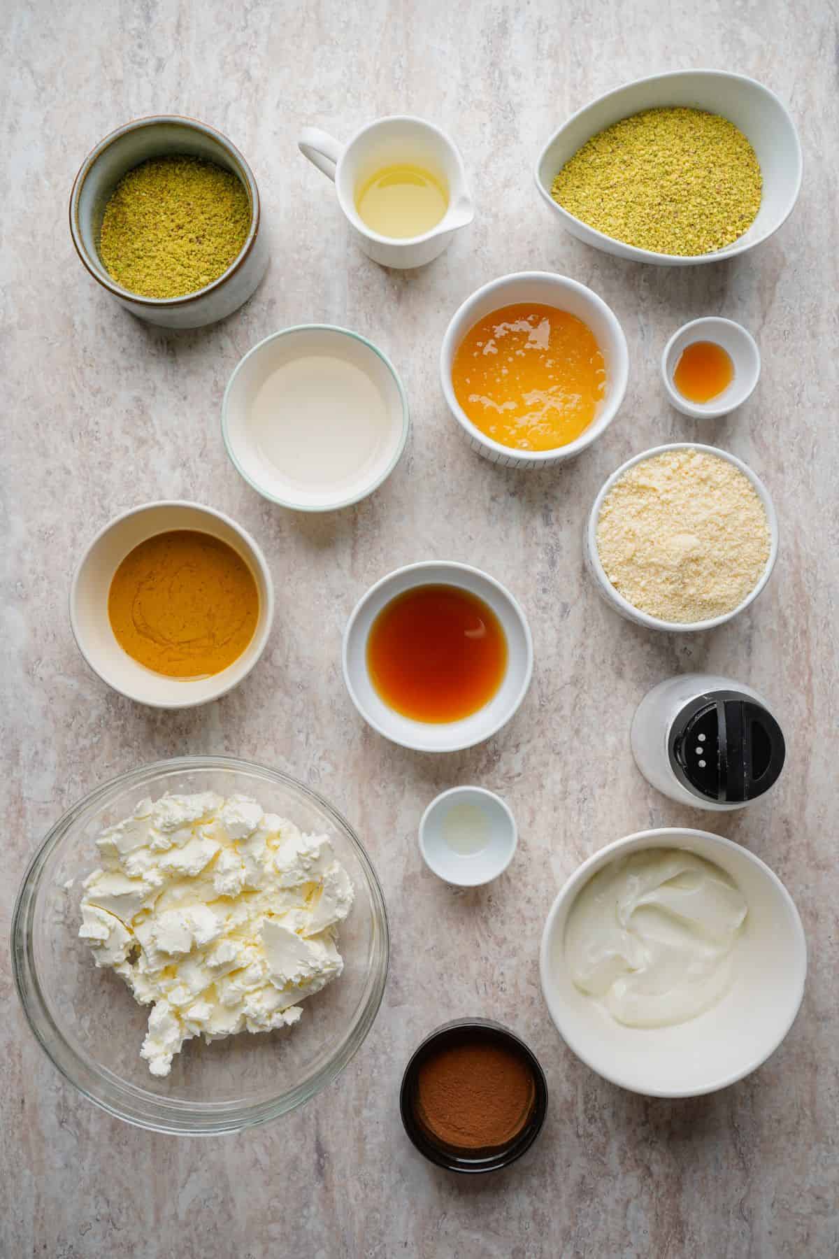 A variety of ingredients for a sweet breakfast including yogurt, honey, pistachios, and spices on a neutral surface.
