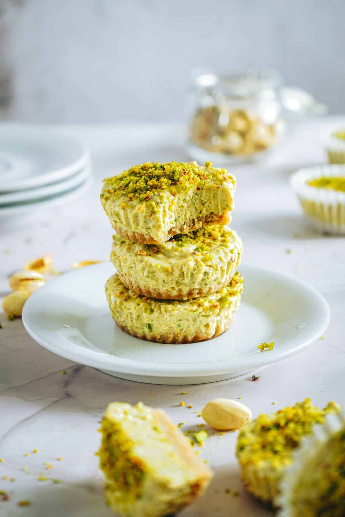 Delicious pistachio cheesecake stacked on a white plate, surrounded by mini cheesecakes and toppings in a bright kitchen setting.