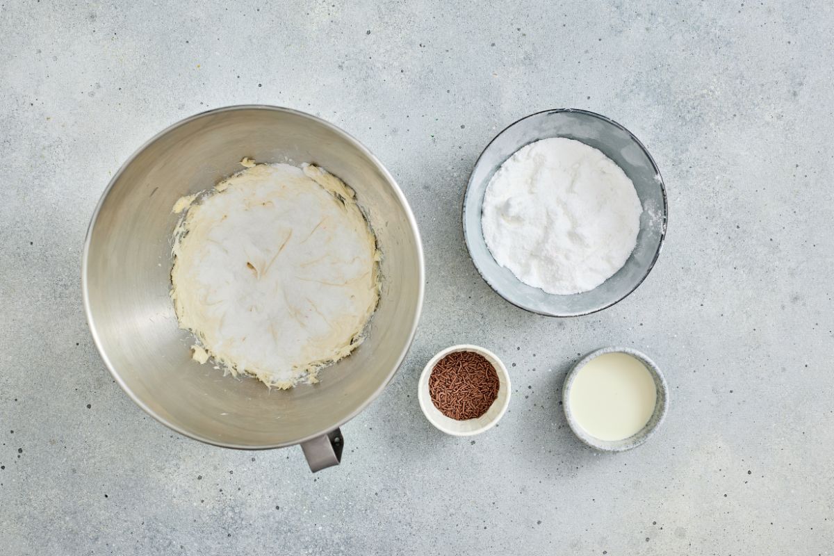 Aerial view of mixing bowls with sugar, flour, melted butter, chocolate sprinkles, and milk on a gray surface.