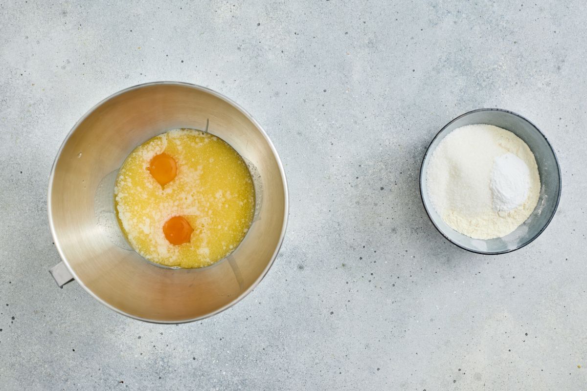 A mixing bowl with eggs and melted butter, and a separate bowl of flour on a light countertop.