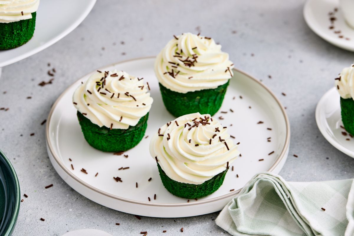 Rich green velvet cupcakes topped with swirls of white frosting and chocolate sprinkles, perfect for celebrations.