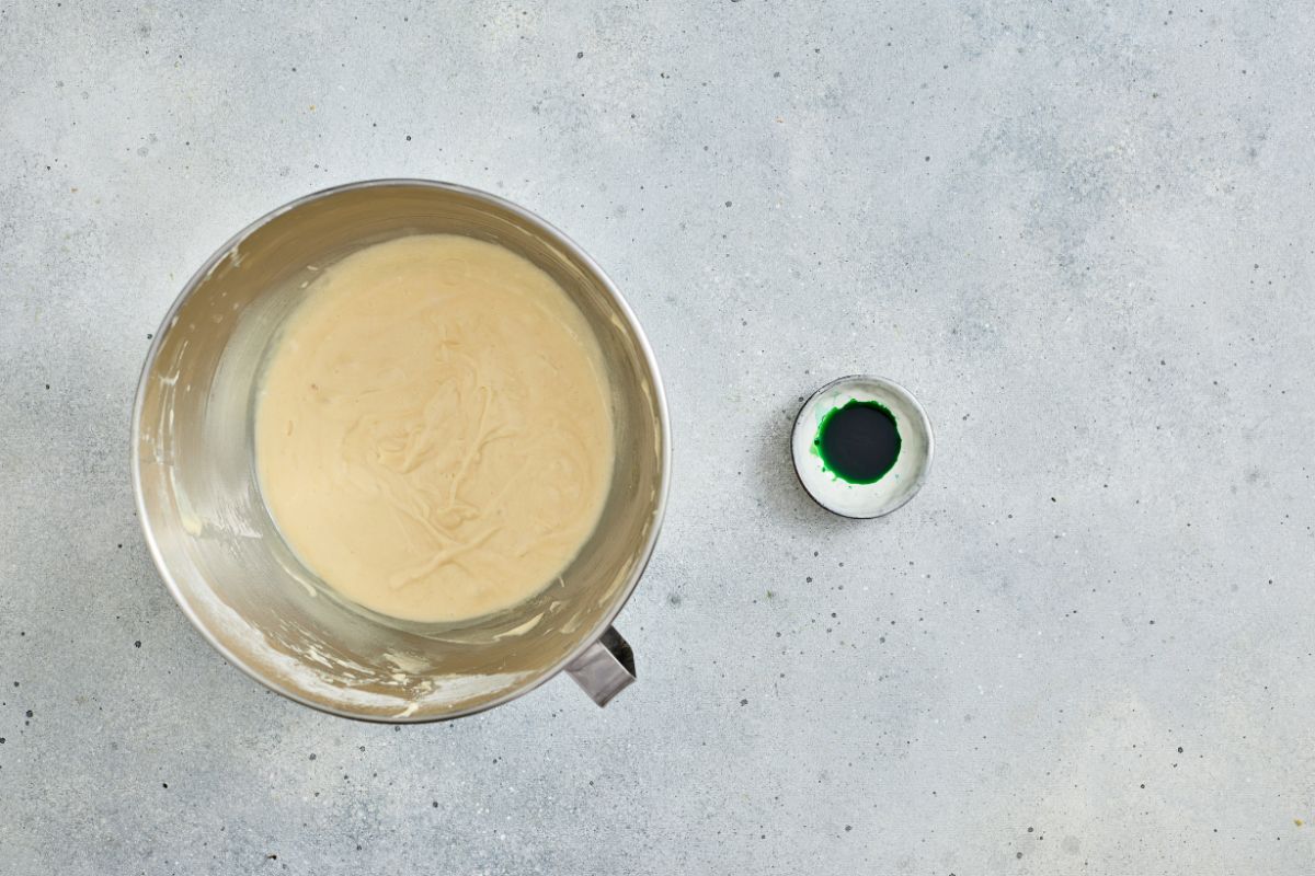 Mixing bowl with cake batter and green food coloring on a gray countertop.