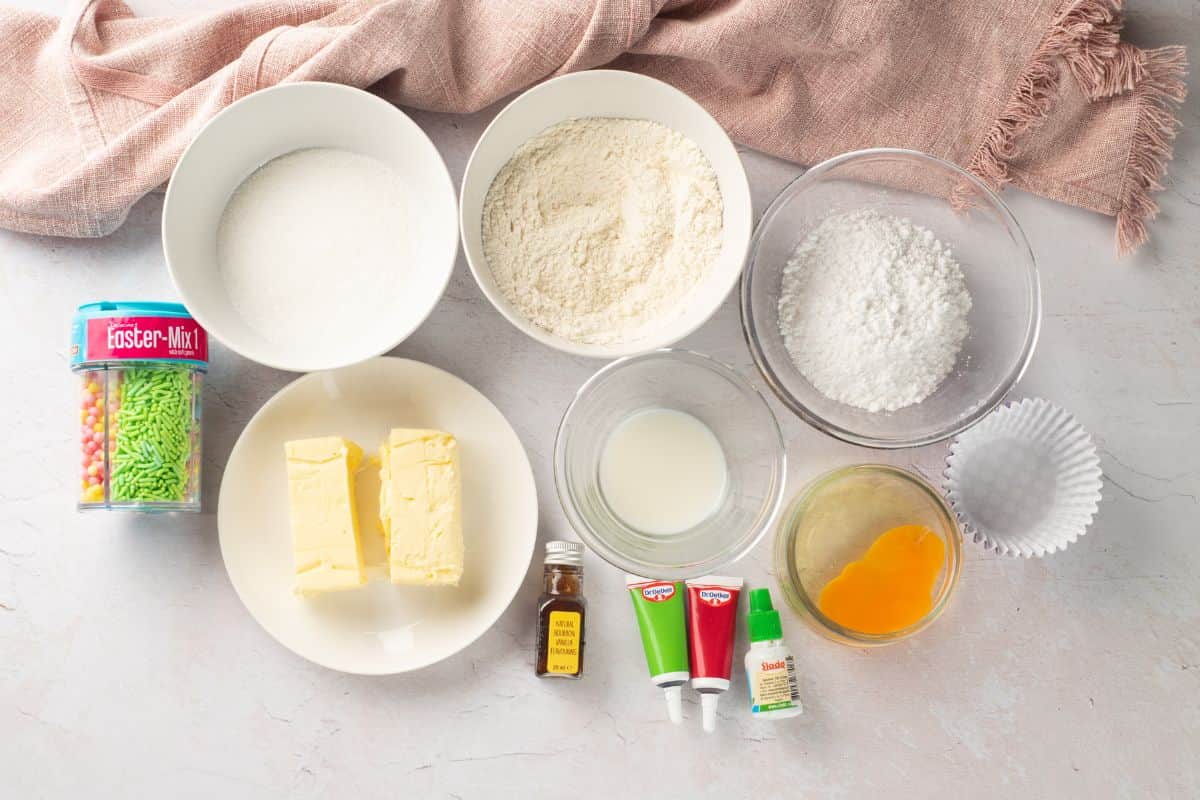 Assorted baking ingredients, such as flour, sugar, butter, eggs, and frosting, arranged on a white surface.