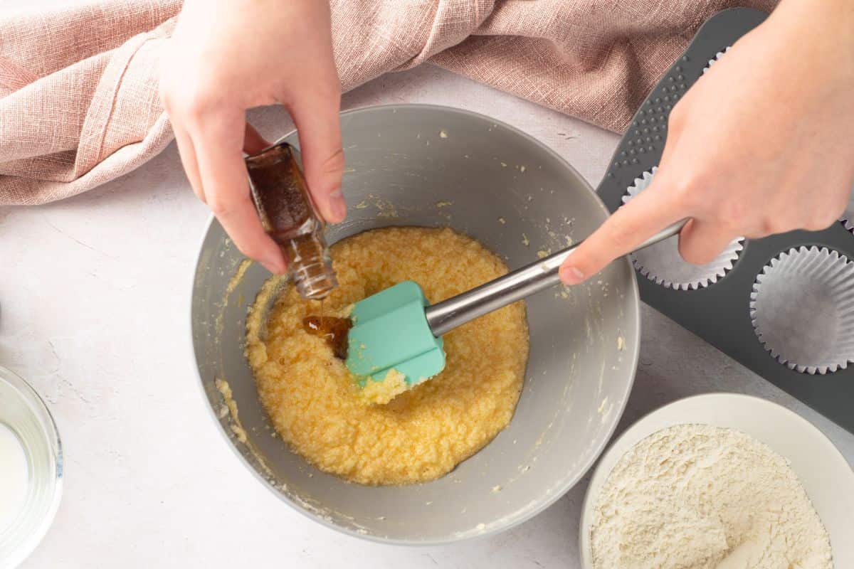 Close-up of hands pouring vanilla extract into a bowl of batter while using a silicone spatula; baking ingredients prep.