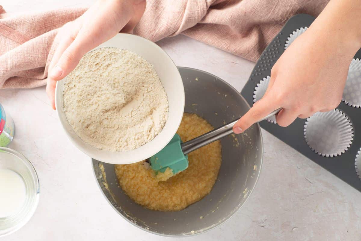 An overhead view of a person pouring flour into a mixing bowl for baking cupcakes, with muffin tins nearby.