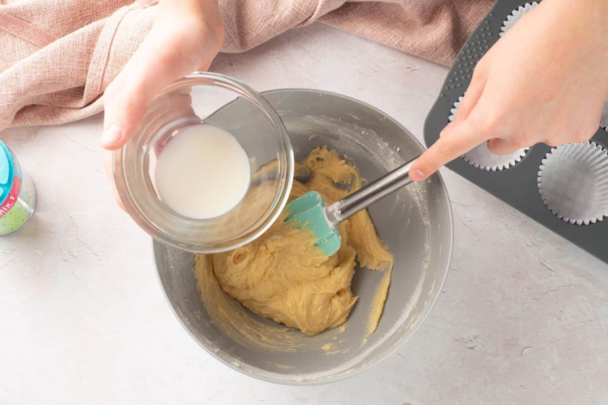 Close-up of hands pouring milk into batter while using a spatula for mixing.