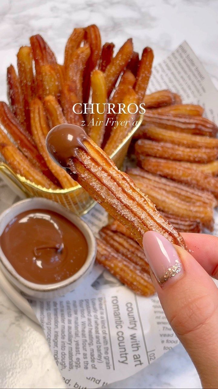 A hand holds up a sugar-coated churro, dipping one end into a bowl of chocolate sauce.