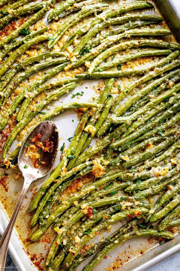 Close up of roasted green beans with melted cheese and herbs on a baking sheet, with a serving spoon resting on the side.