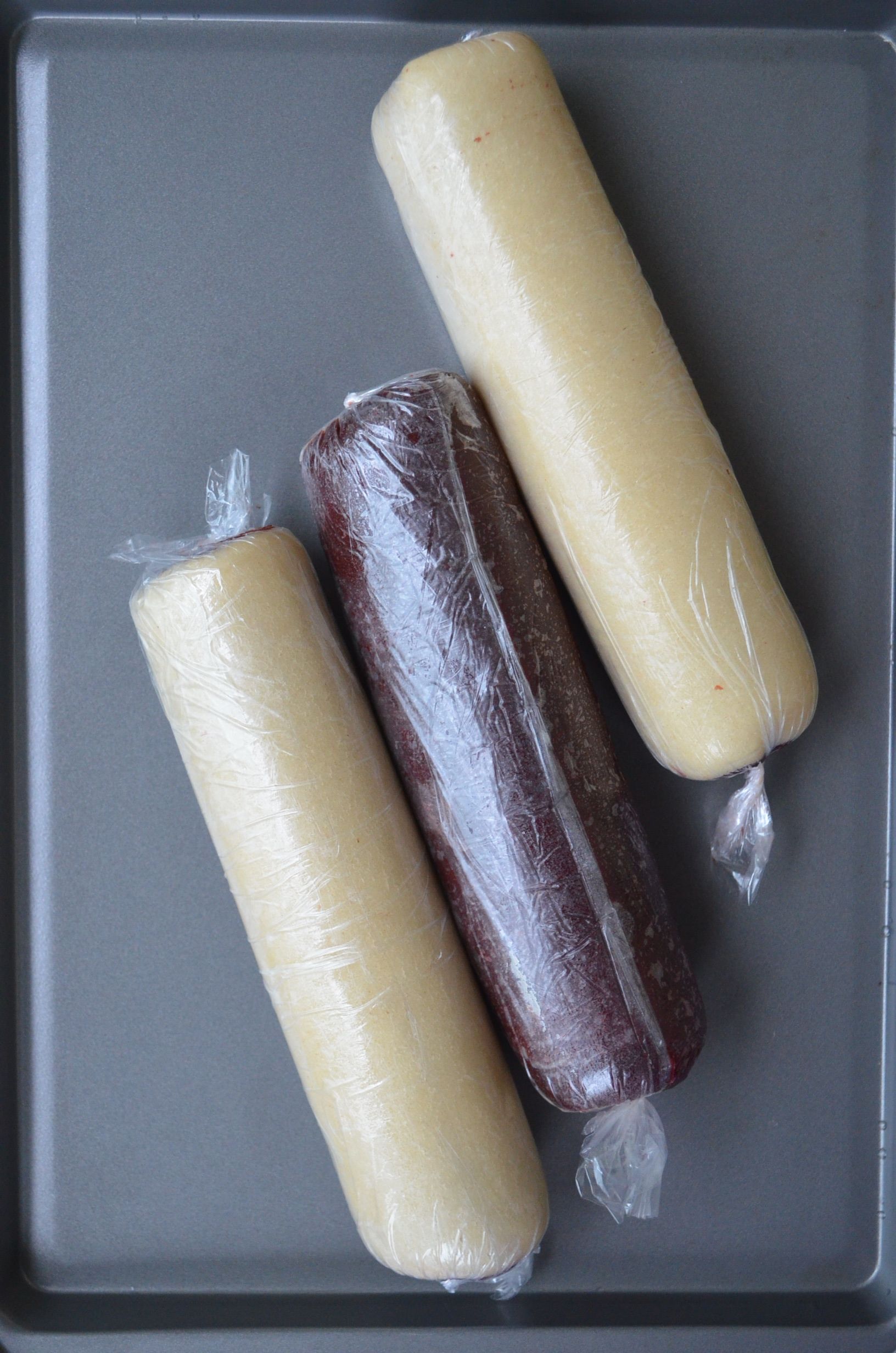 Three rolls of cookie dough, two light-colored and one dark, wrapped in plastic wrap on a baking sheet.
