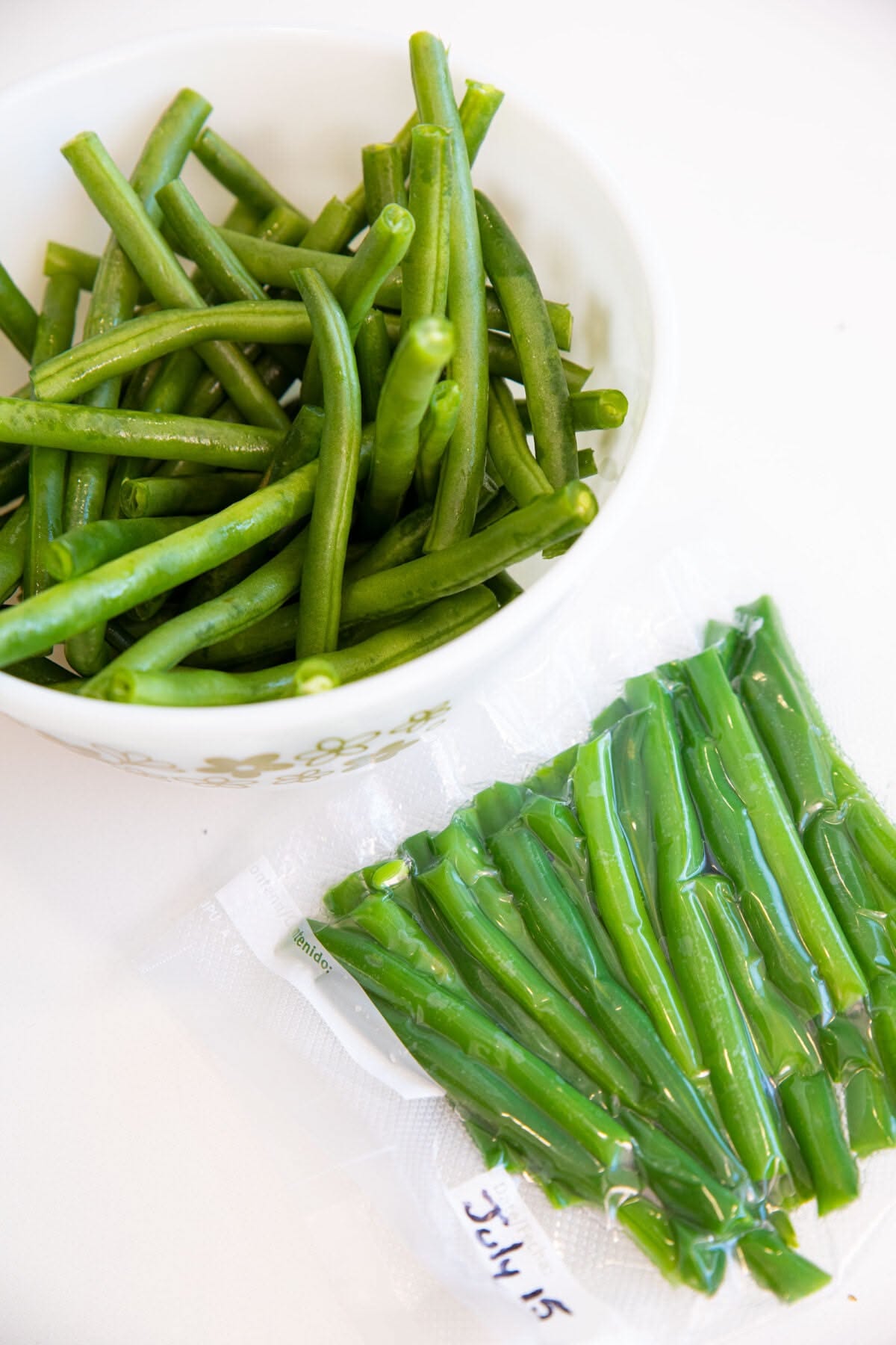 A white bowl holds blanched green beans, next to a vacuum-sealed bag of frozen green beans labeled 'July 15'.