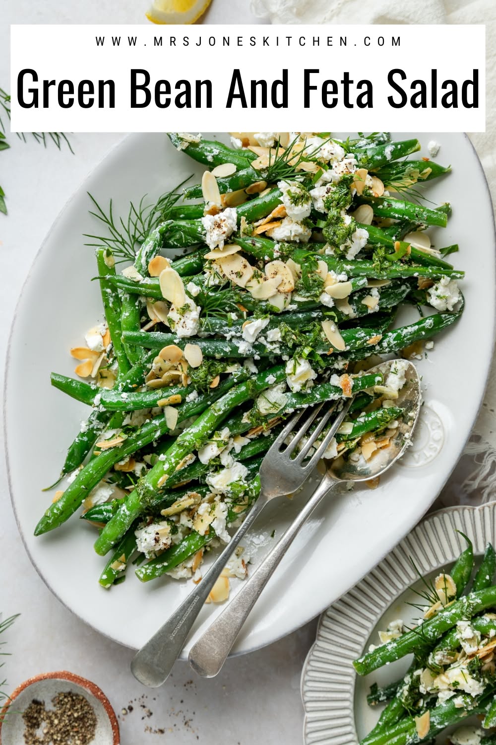 White oval platter filled with cooked green beans, crumbled feta, sliced almonds, and fresh herbs, with a serving fork and…