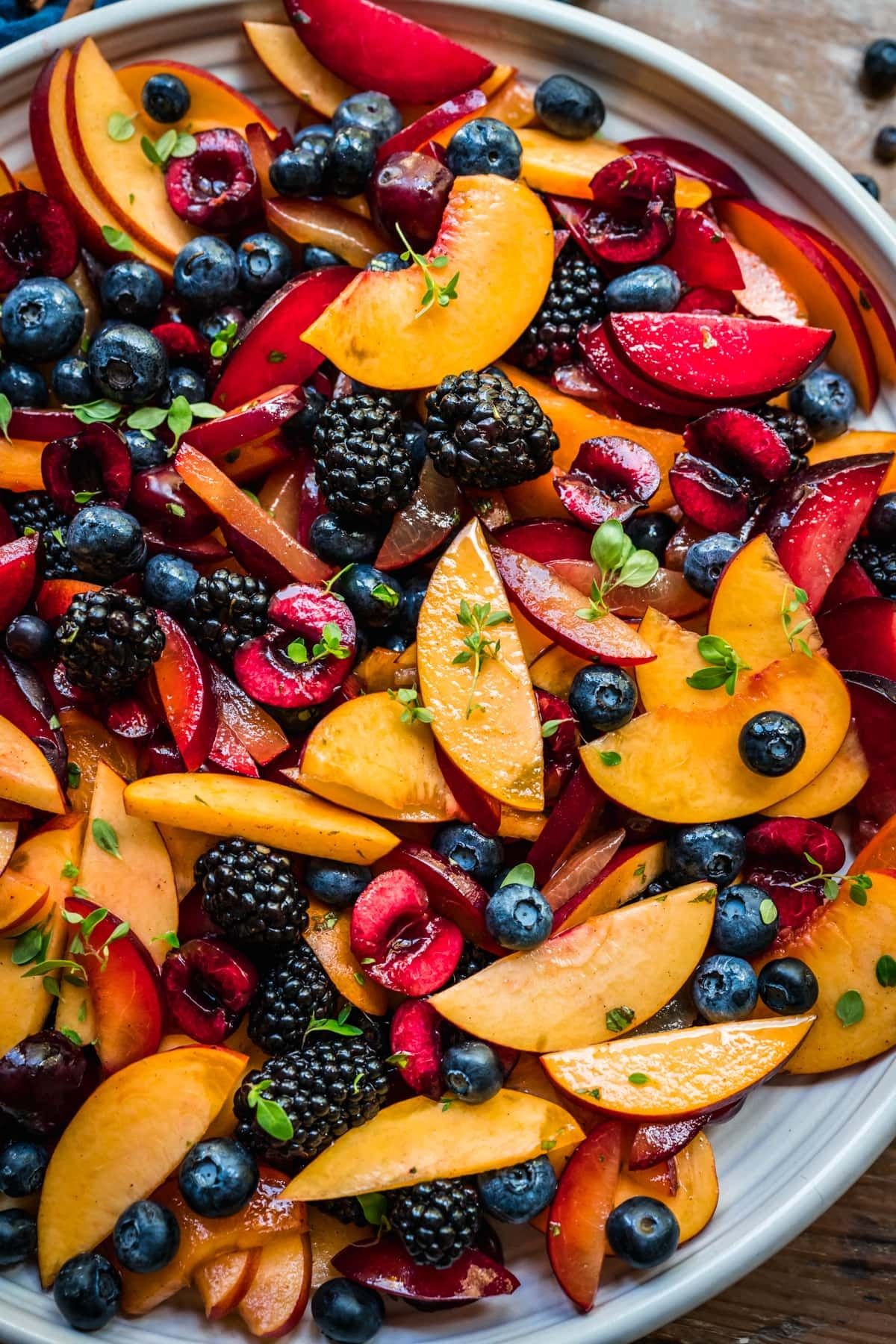 Close-up of a salad with sliced peaches, plums, blueberries, blackberries, and halved cherries, garnished with fresh thyme…