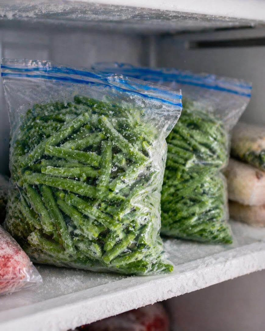 Frozen cut green beans in clear freezer bags, stored on a frost-covered freezer shelf.