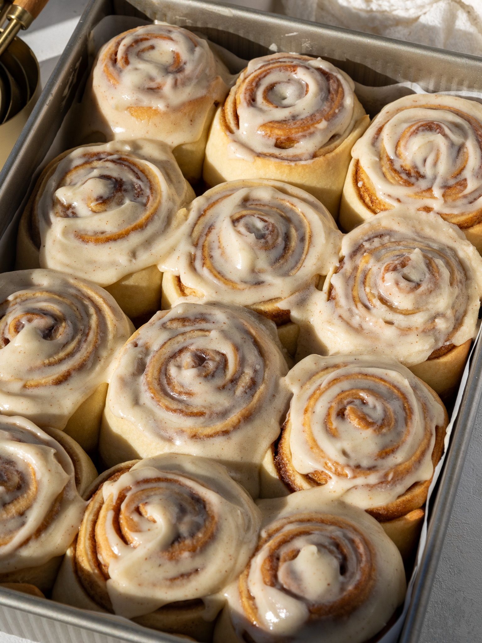 Close-up of a baking pan filled with frosted cinnamon rolls, each swirled with cinnamon sugar and topped with a generous…