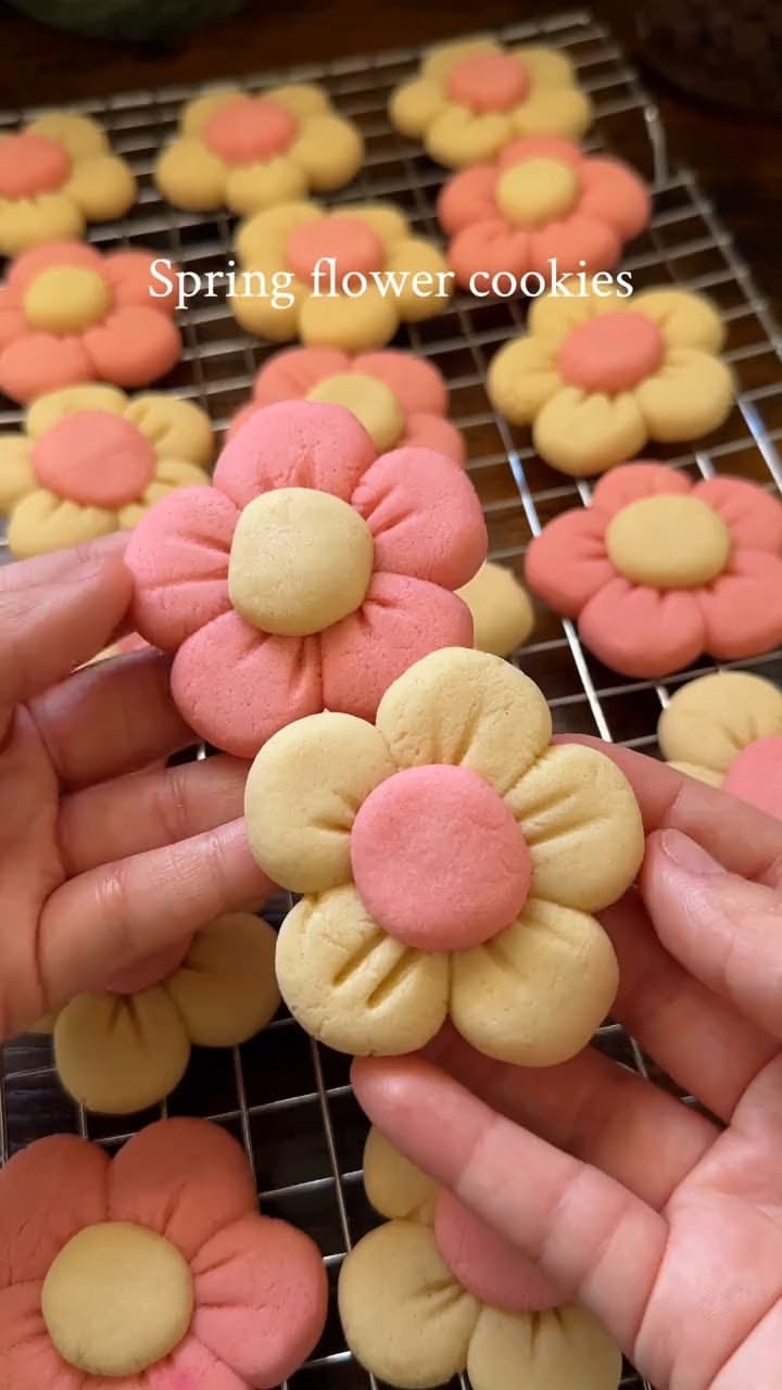 Close-up of hands holding two flower-shaped cookies, with several more on a wire rack in the background.