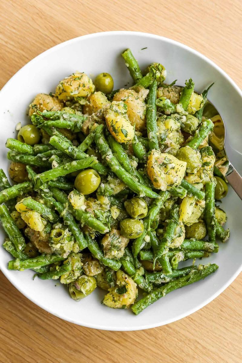 Warm green bean and potato salad with green olives and dill in a white bowl with a spoon.