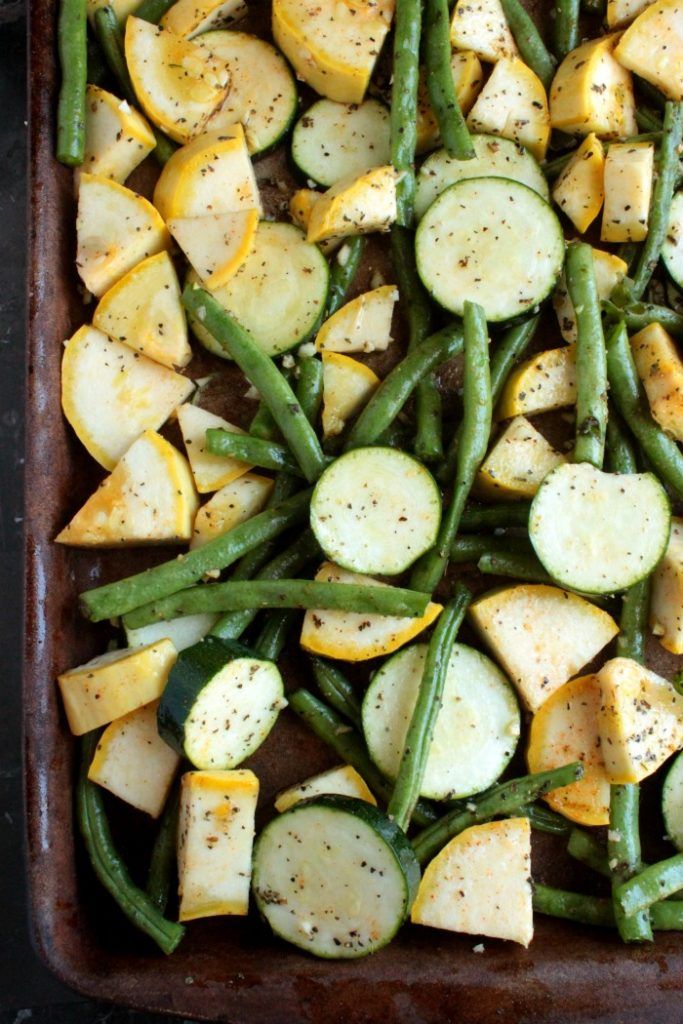 Close up of green beans, sliced zucchini, and sliced yellow squash seasoned with herbs on a baking sheet