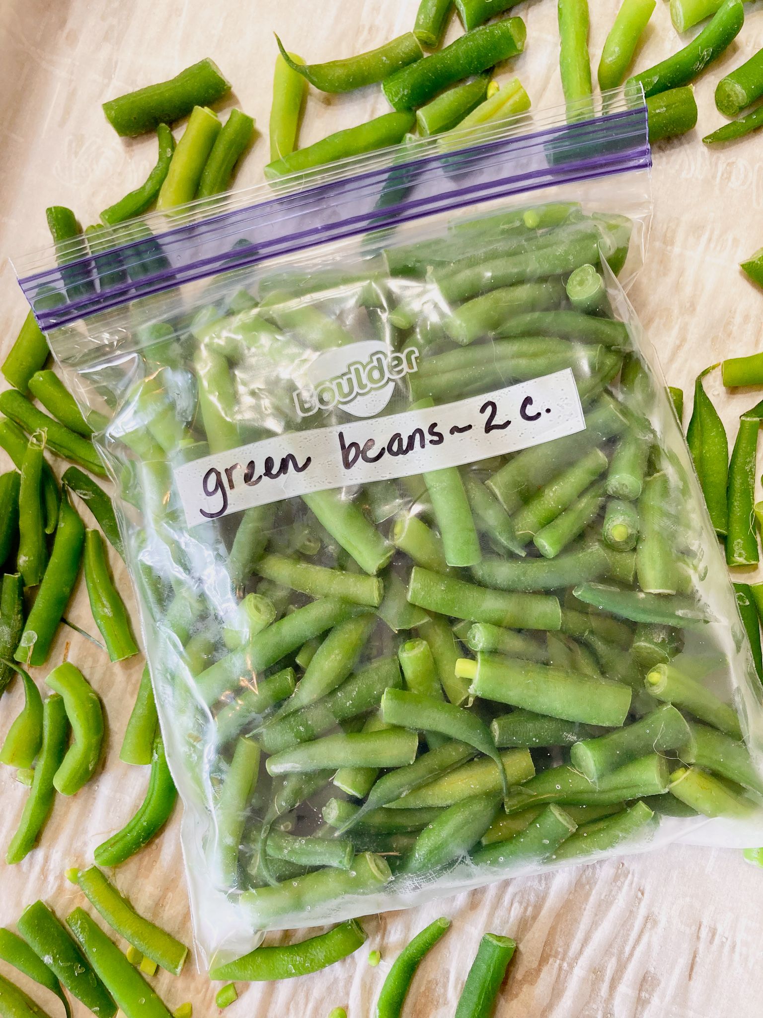 A plastic freezer bag filled with cut, frozen green beans sits on a surface covered with more individual frozen green beans.