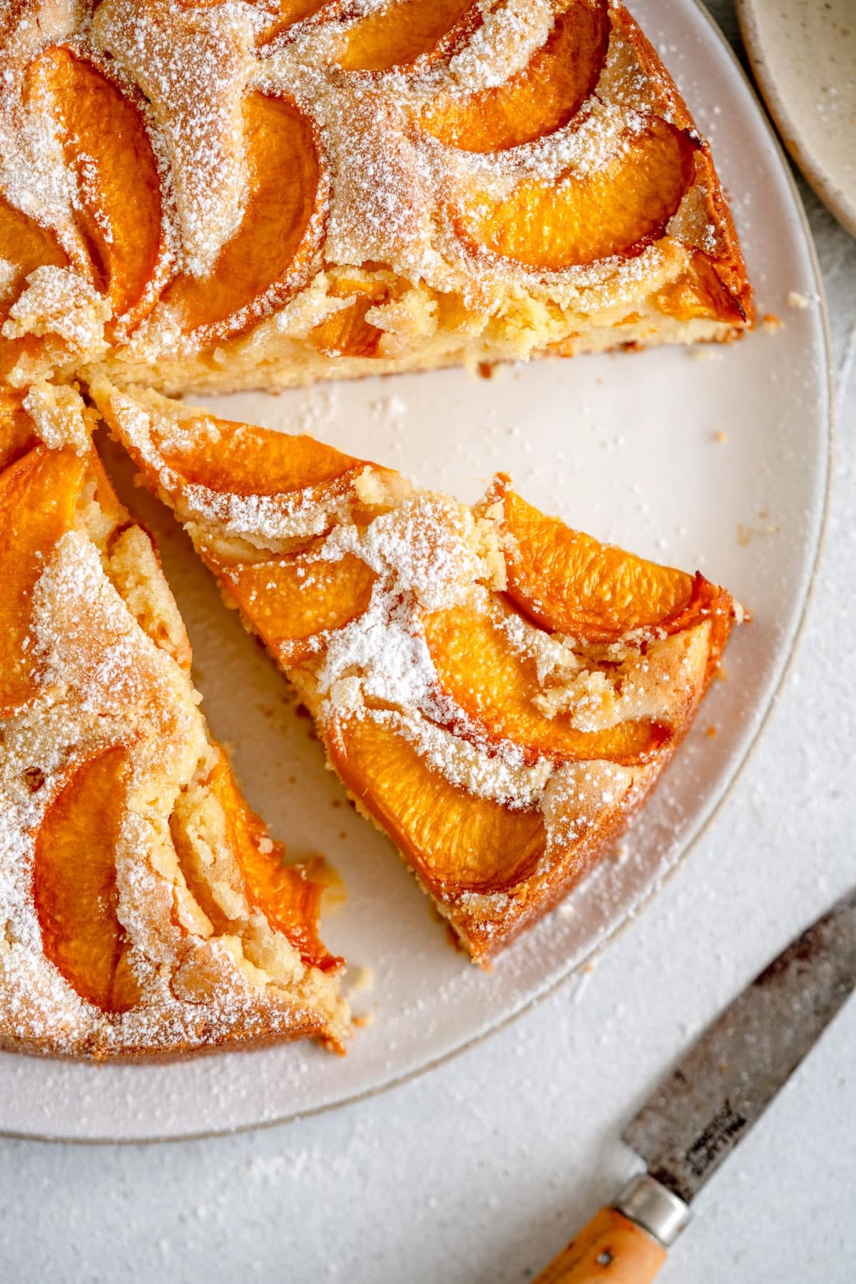 A round, sliced peach cake covered in powdered sugar on a white plate, with a vintage-style knife resting next to it.