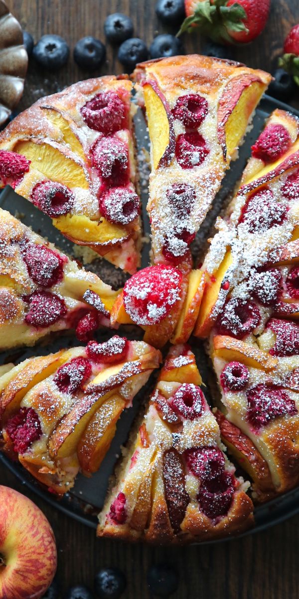 Slices of cake topped with fresh peach slices, raspberries, and powdered sugar, on a dark plate.