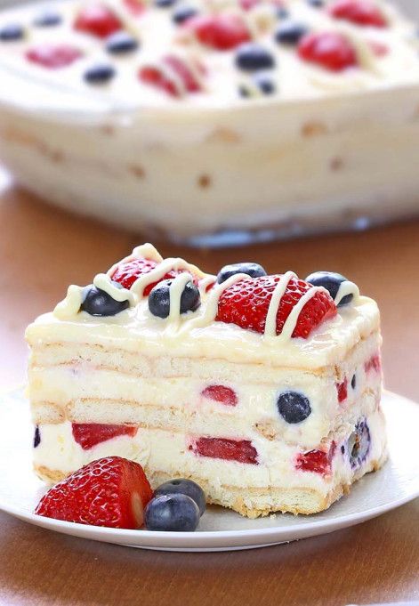 Close-up of a slice of no-bake cake with layers of cream and cookies, studded with blueberries and strawberries, topped with…