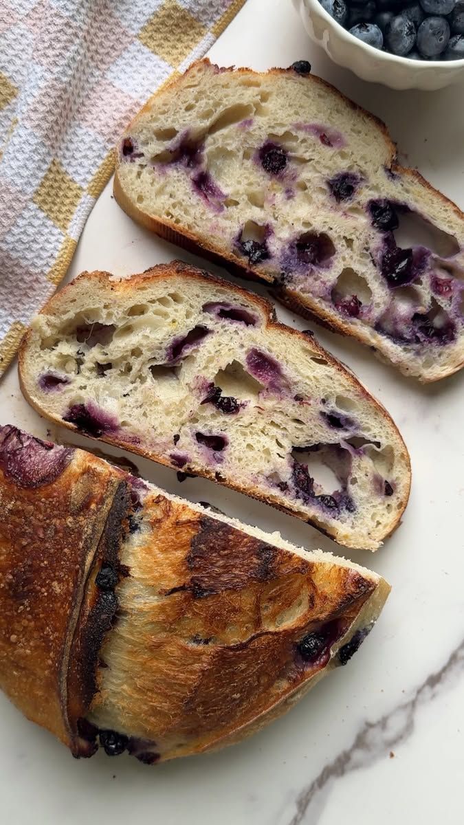 Close-up of three slices of sourdough bread studded with blueberries, with a small bowl of blueberries in the background.
