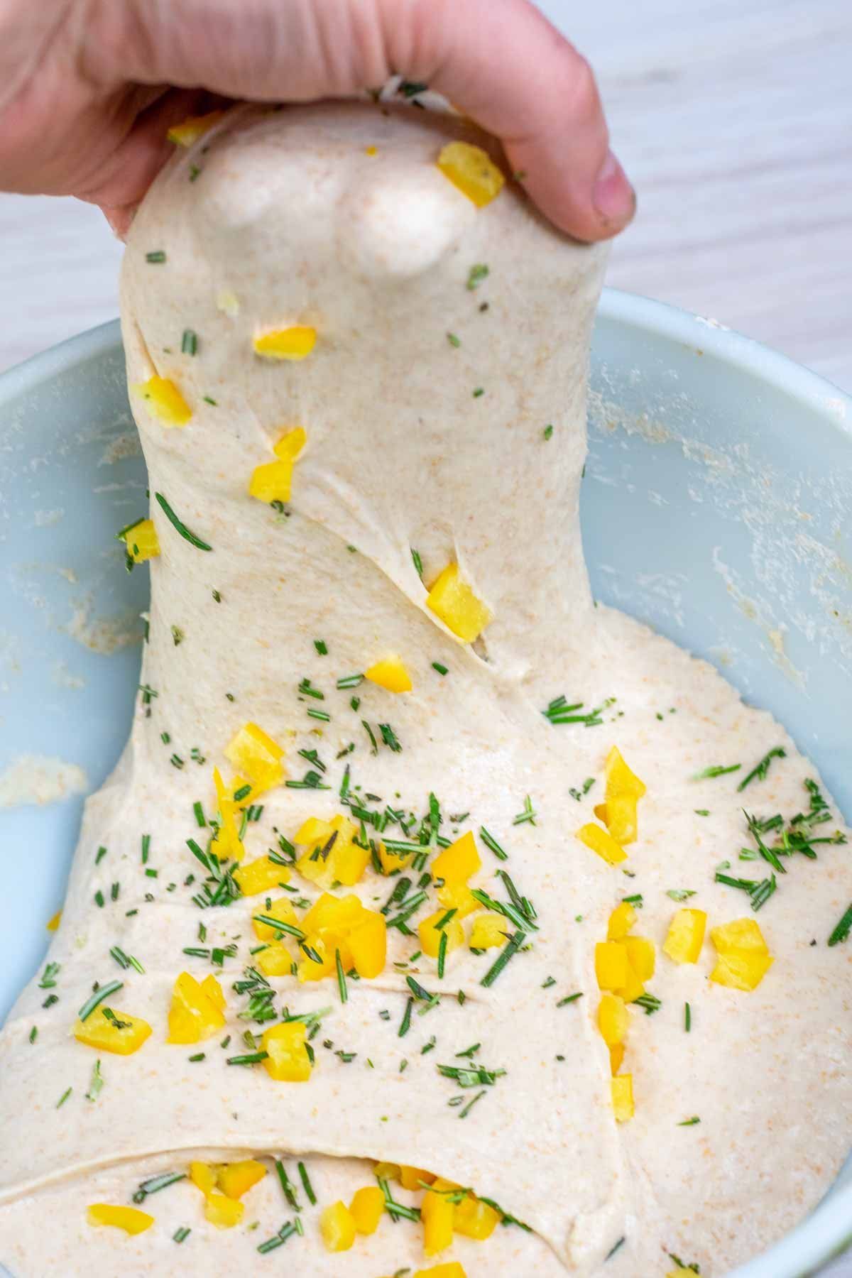 A person's hand stretching a pale-colored sourdough dough out of a light blue bowl, revealing small yellow pieces of…