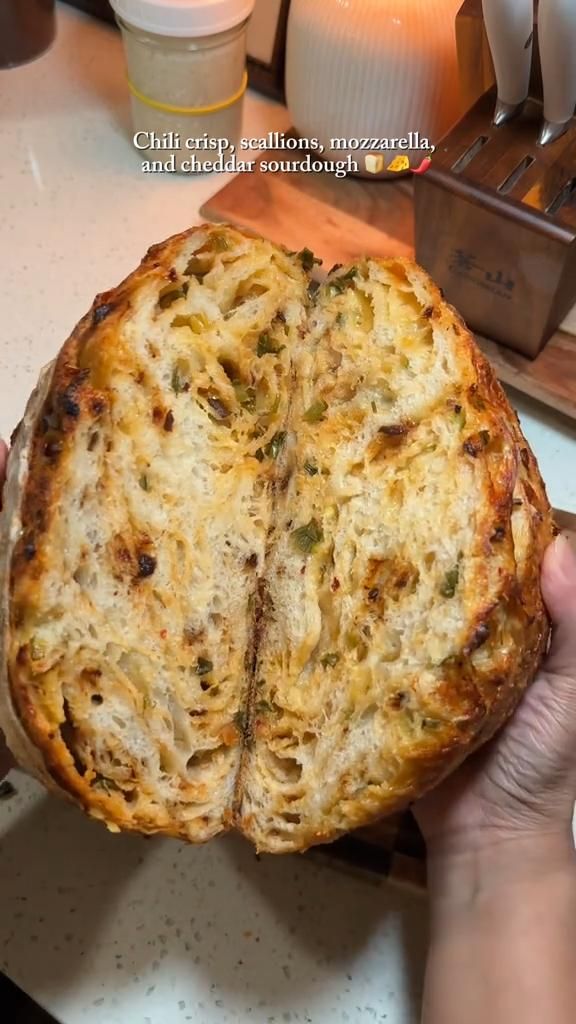 A round loaf of sourdough bread, split open to reveal a soft, airy interior speckled with green scallions and melted cheese.