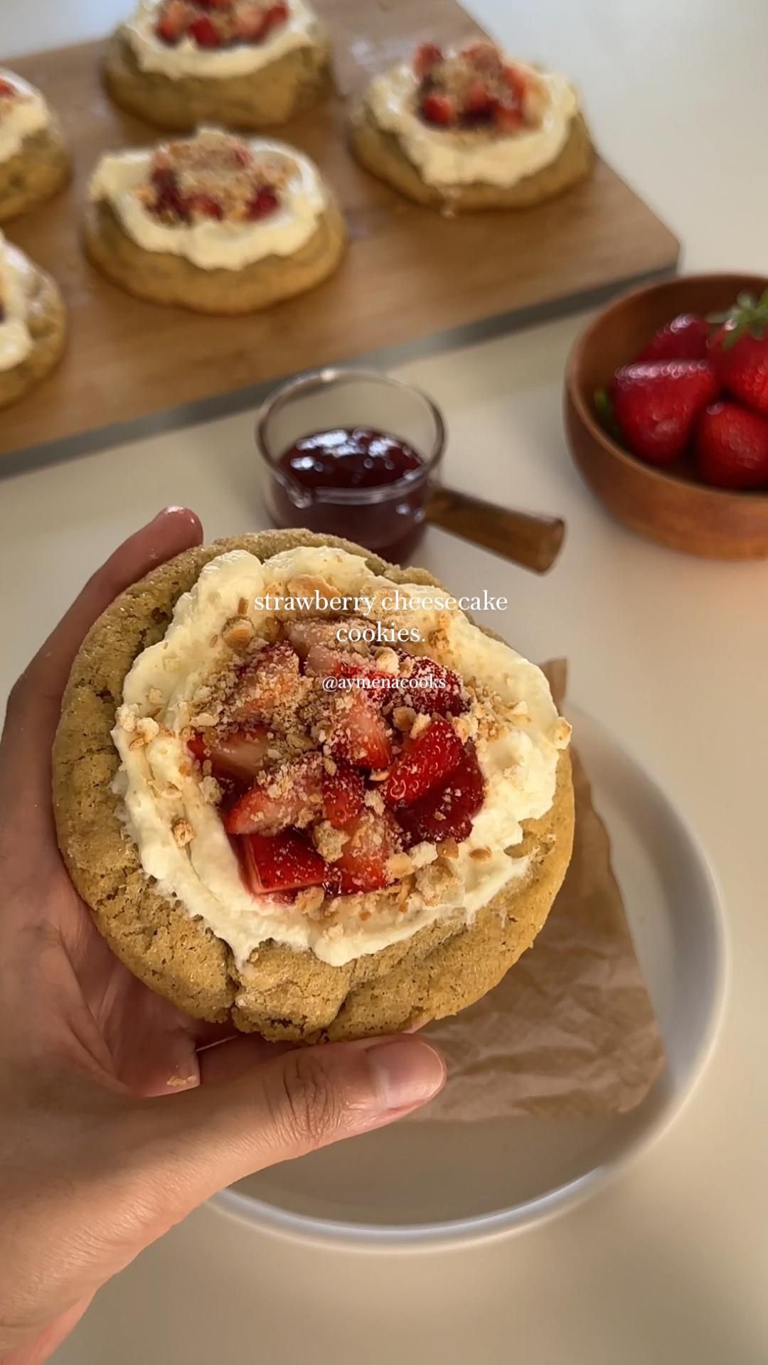 Hand holding a large, round cookie topped with white frosting, diced strawberries, and crumbled topping.