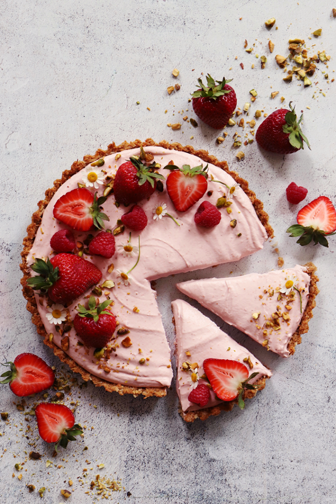 Round strawberry mousse tart with two slices removed, garnished with whole and halved strawberries, raspberries, small white…