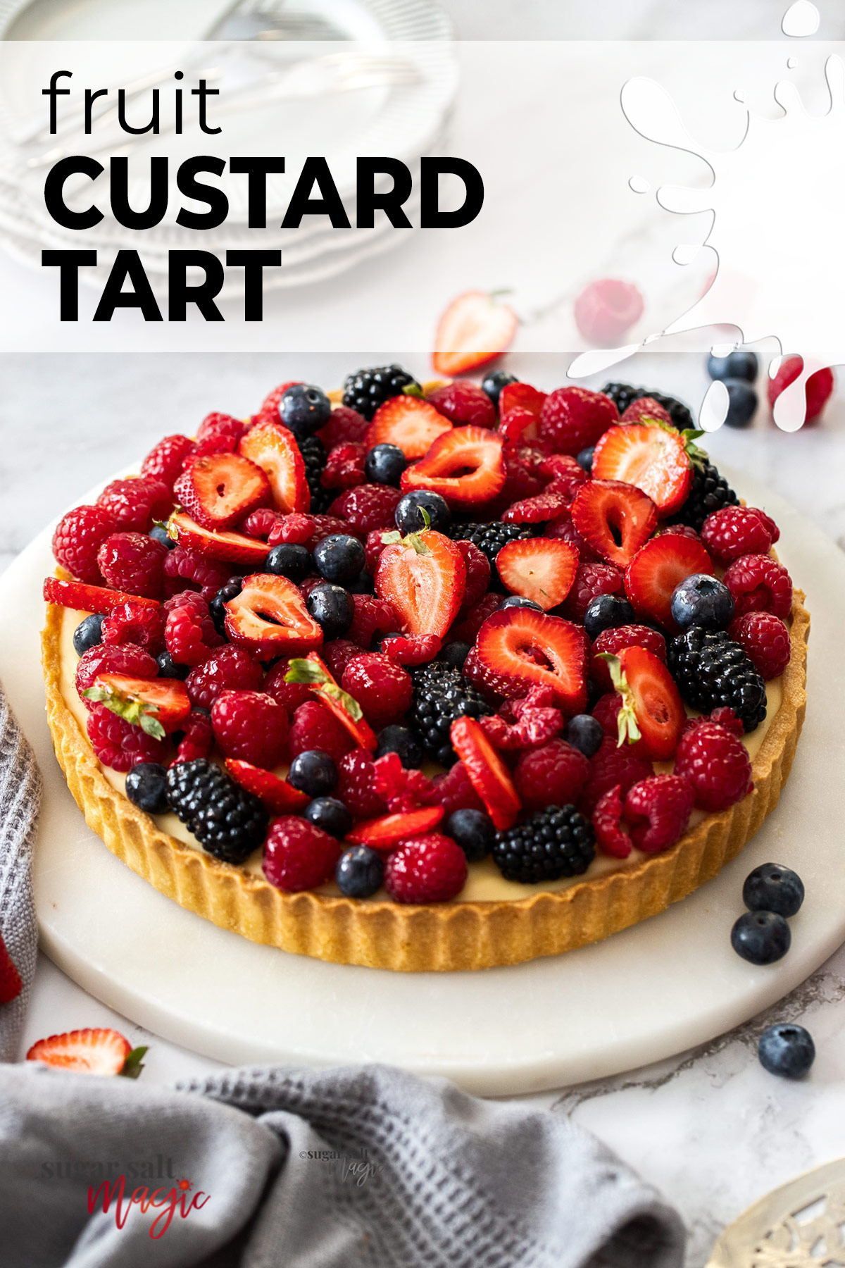 A round fruit custard tart topped with whole and sliced red raspberries, blueberries, blackberries, and strawberries, on a…