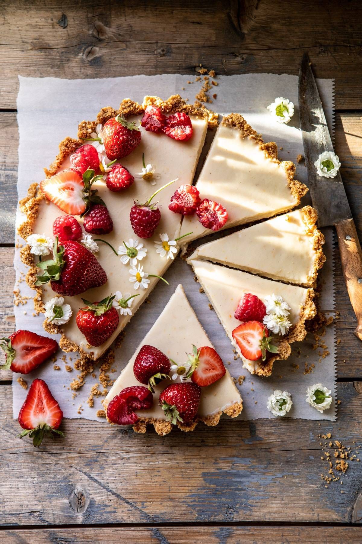 Lemon tart with a crumb crust, garnished with fresh strawberries, raspberries, and small white flowers, with several slices…