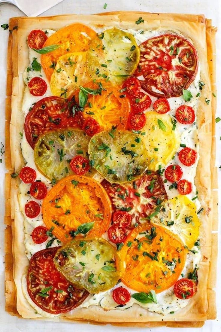 Close up, overhead view of a savory tart topped with sliced heirloom tomatoes, halved cherry tomatoes, and herbs on a phyllo…