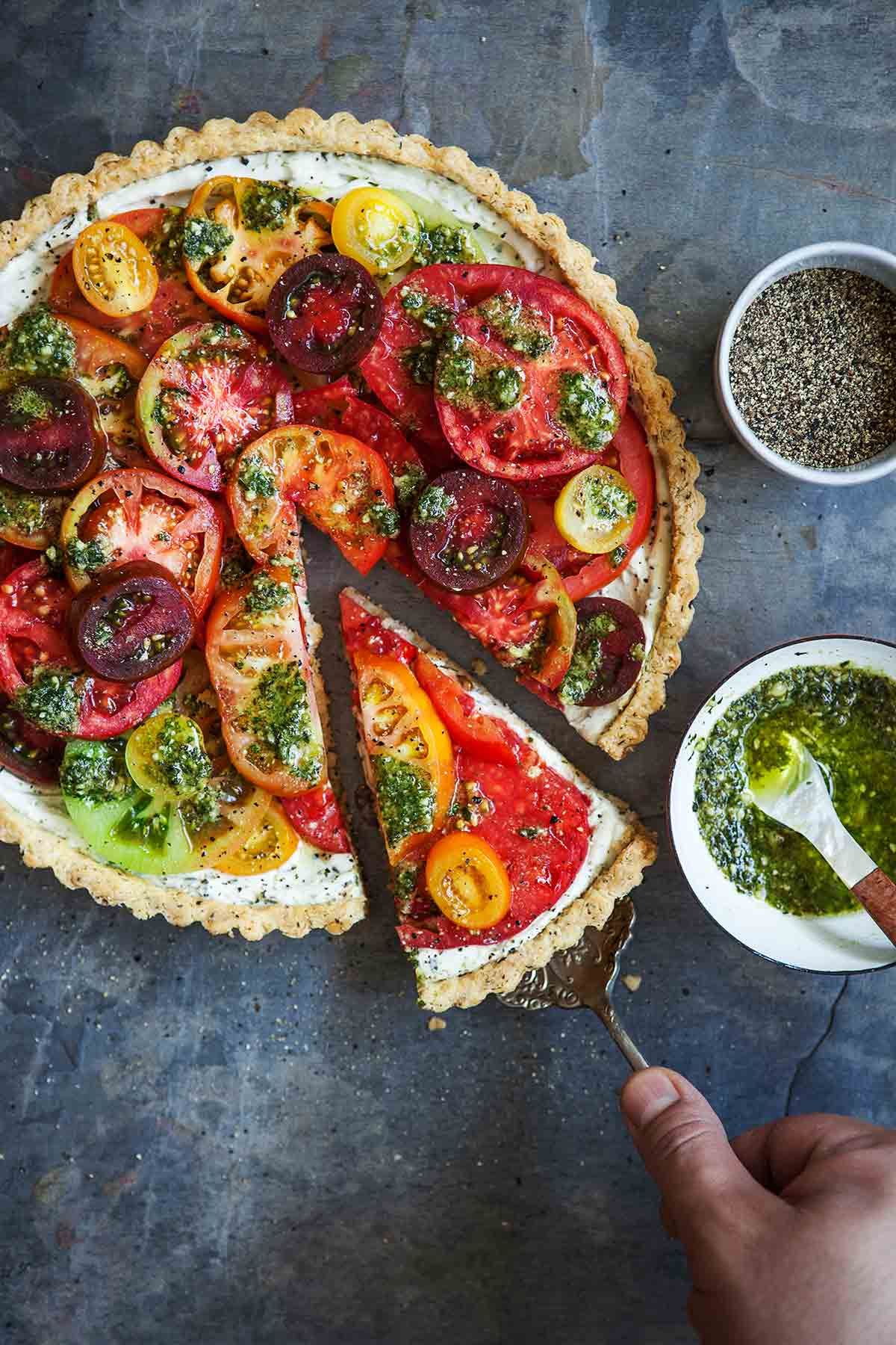 A round tomato tart with colorful sliced tomatoes and green pesto on top. A slice is being removed with a server.