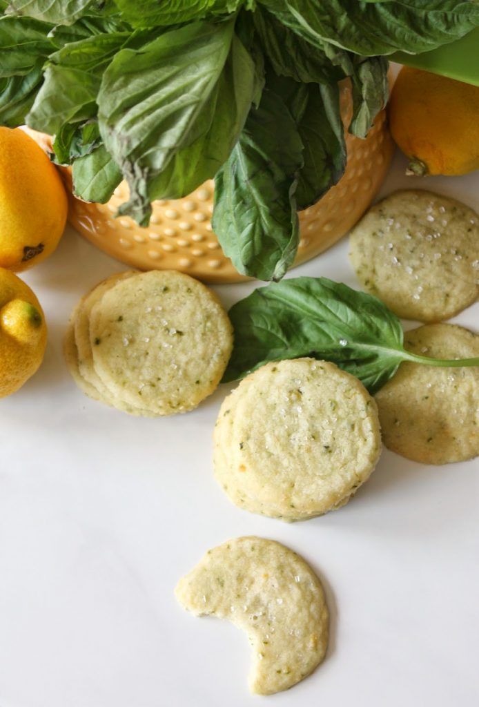 Round, pale yellow cookies with green flecks and a sugar topping arranged on a white surface with fresh lemons and basil.