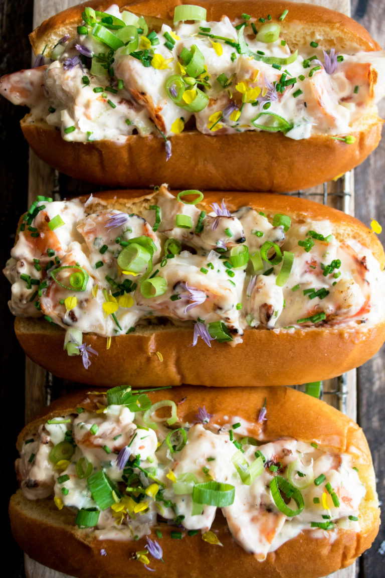 Three creamy shrimp rolls on buttered split-top buns, garnished with chives and small yellow and purple edible flowers.