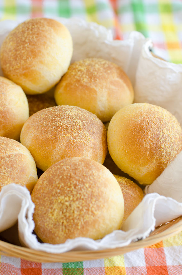 A basket filled with golden-brown cornbread rolls dusted with cornmeal, set on a colorful plaid tablecloth.