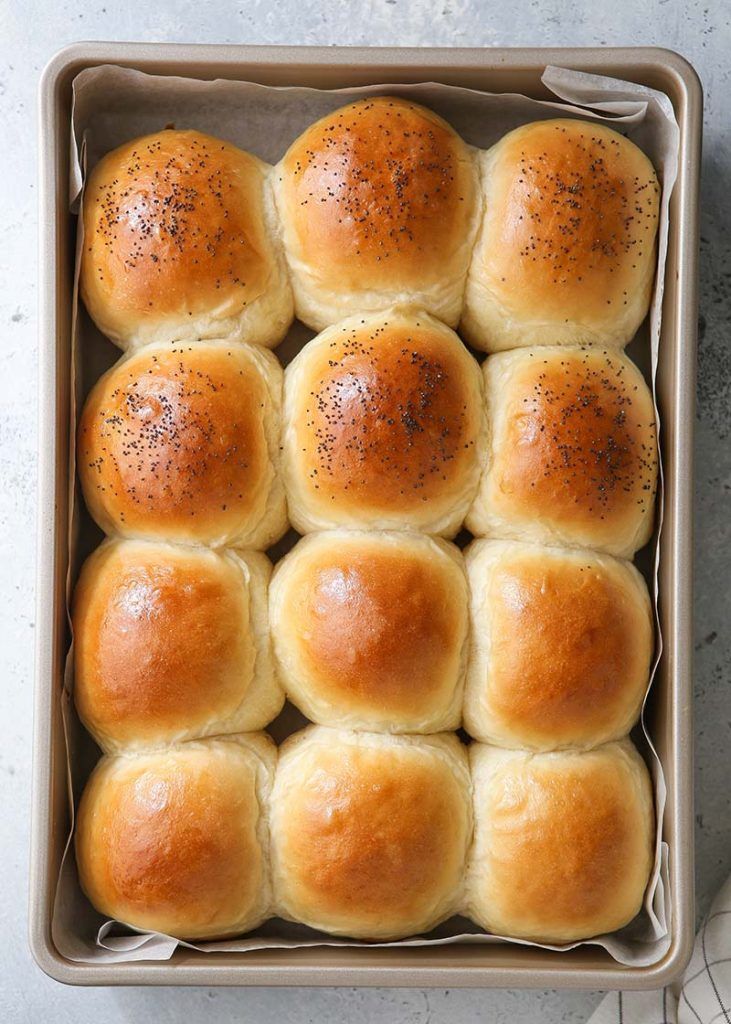 Newly baked fluffy slider buns, some topped with poppy seeds, arranged in rows inside a baking pan.