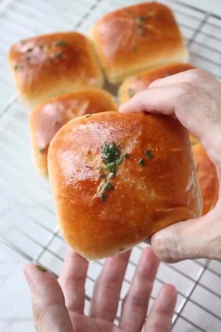 Hand holding a square, golden brown bun with green herb topping, with other buns on a wire rack in the background