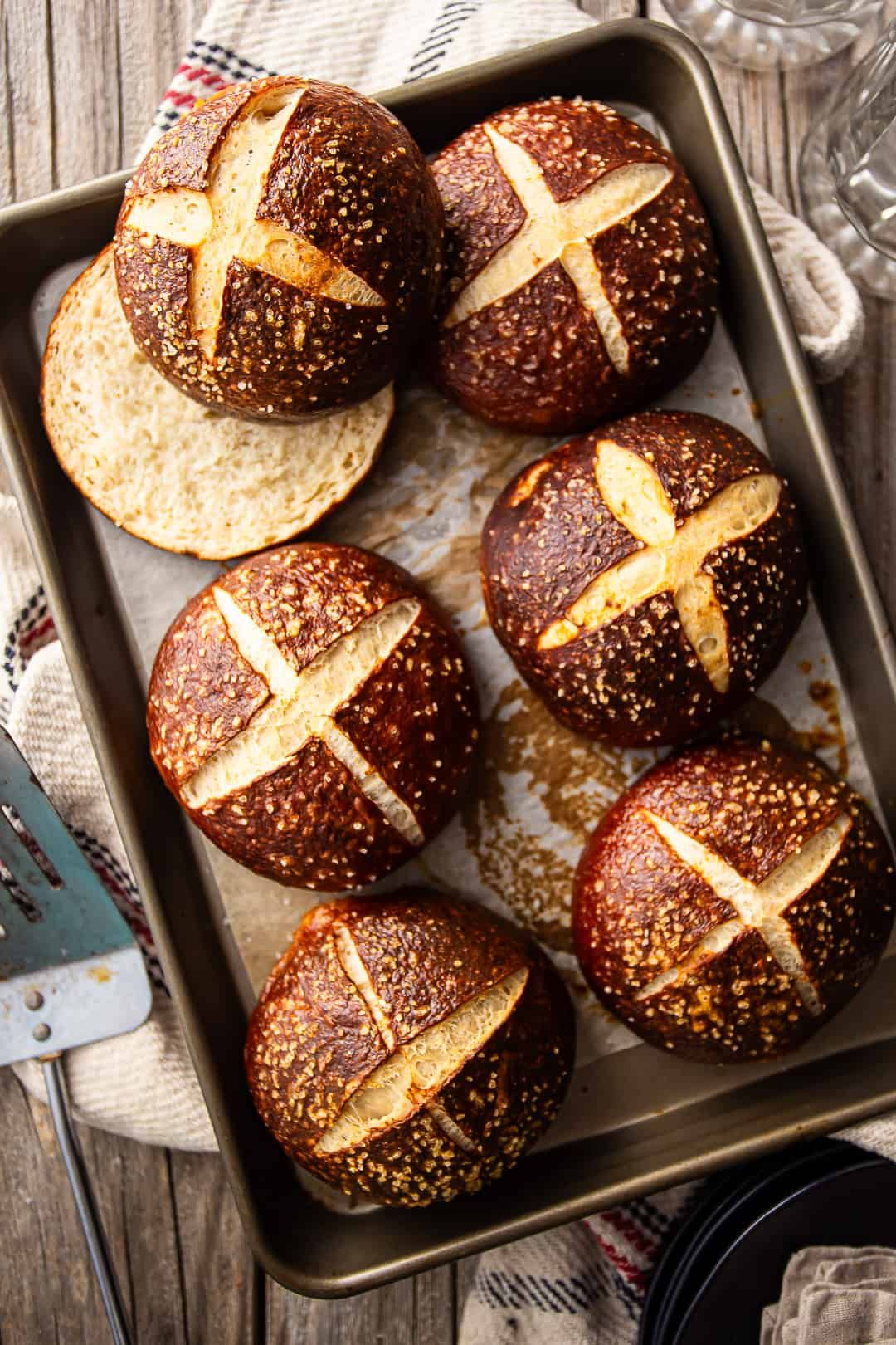 Several freshly baked pretzel buns with scored tops and a dusting of salt on a baking sheet, with one bun sliced open.