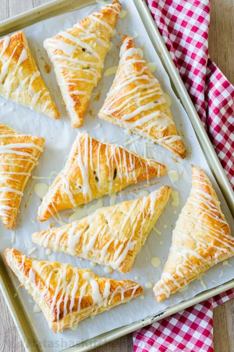 Baked apple turnovers with a white sugar glaze drizzled over them, on a parchment-lined baking sheet.