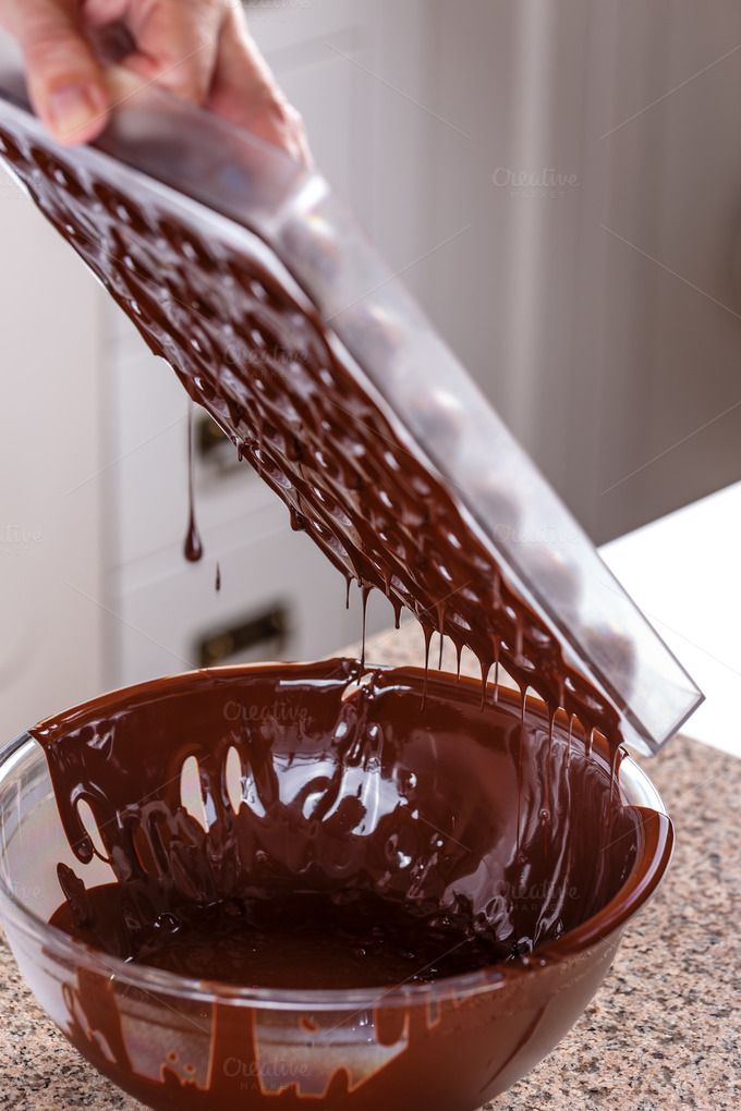 A person pouring excess melted chocolate from a clear plastic mold back into a glass bowl, with drips of chocolate falling.