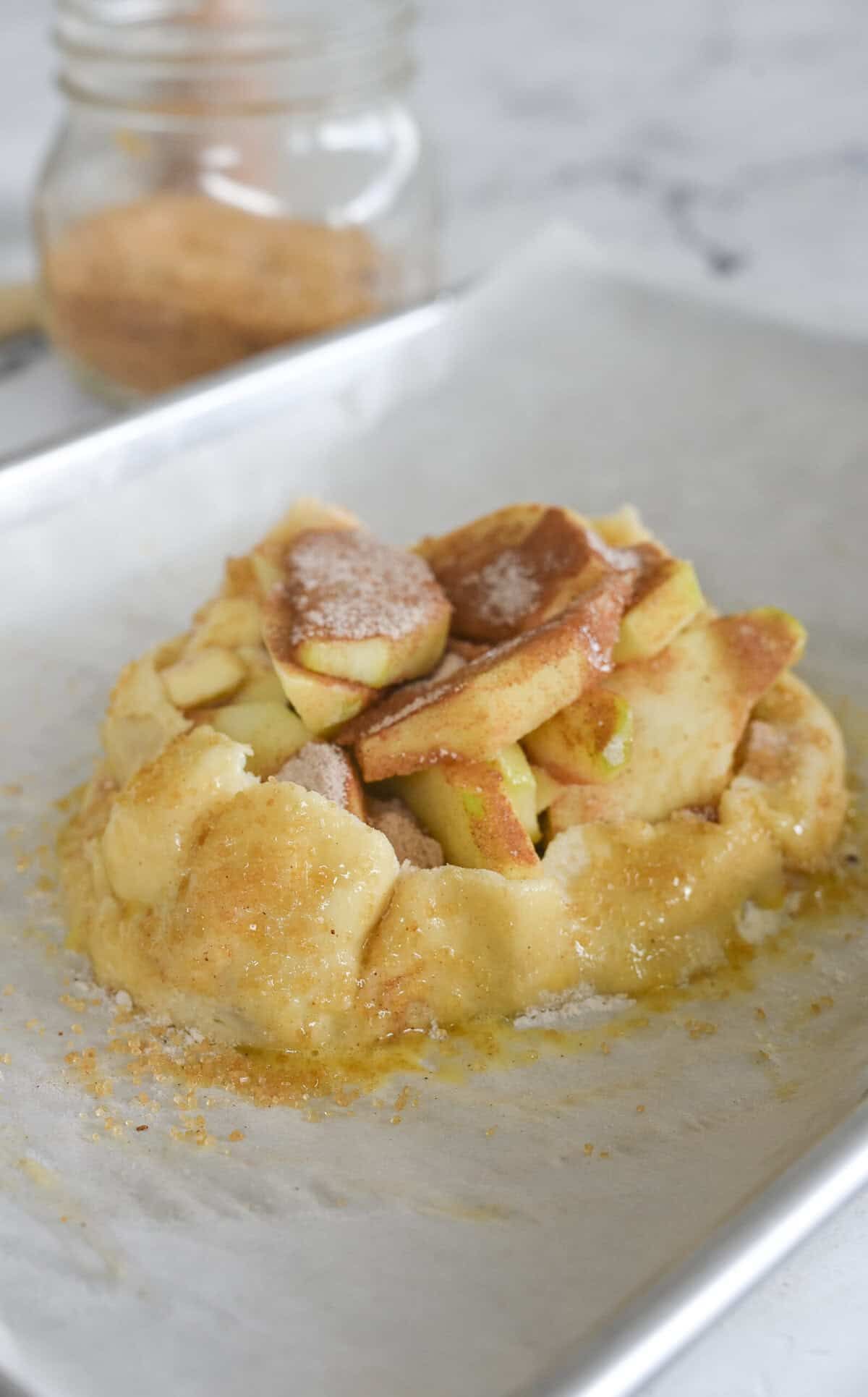 Unbaked individual apple galette on parchment paper, with pie crust folded around apple slices and sprinkled with cinnamon…
