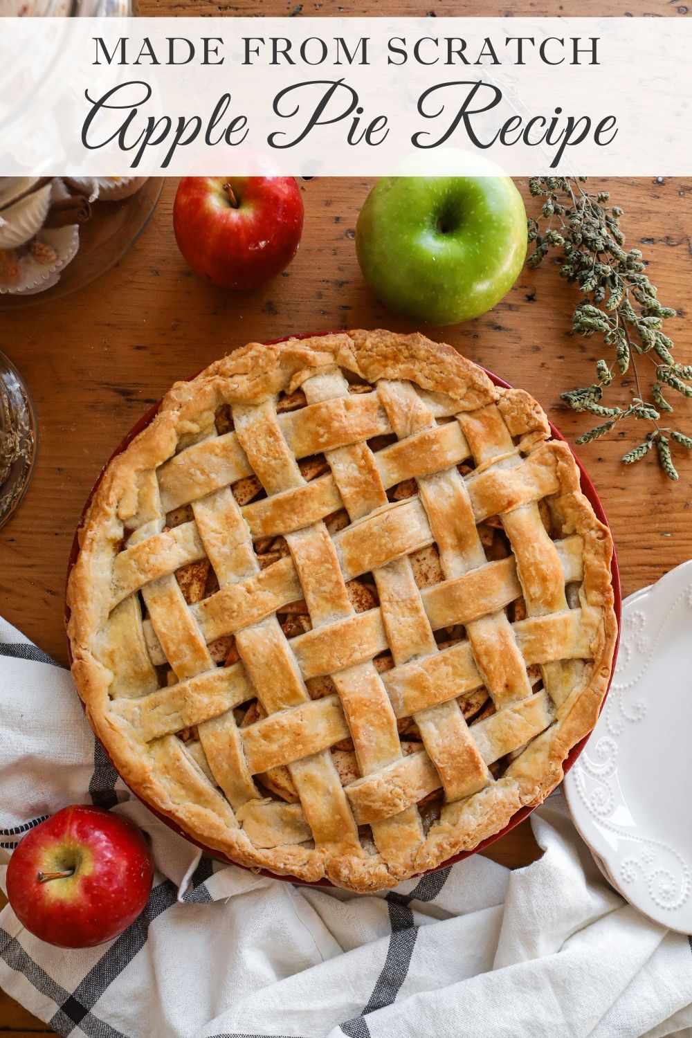 Baked apple pie in a red dish with a golden-brown lattice crust, surrounded by fresh apples on a wooden table.